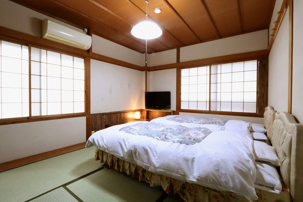 Una habitación japonesa tradicional con suelo de tatami, una cama con colcha floral, un televisor sobre un soporte de madera y puertas correderas shoji.