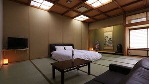 Ein traditioneller japanischer Raum mit Tatami-Boden, einem Bett mit weißer Bettwäsche, einem dunklen Holz-Couchtisch, einer schwarzen Couch, einem Fernseher auf einem Standfuß und einem großen Fenster mit Jalousien.