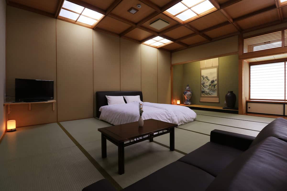 Una habitación tradicional japonesa con suelo de tatami, una cama con ropa de cama blanca, una mesa de centro de madera oscura, un sofá negro, un televisor sobre un mueble bajo y un ventanal con persianas.