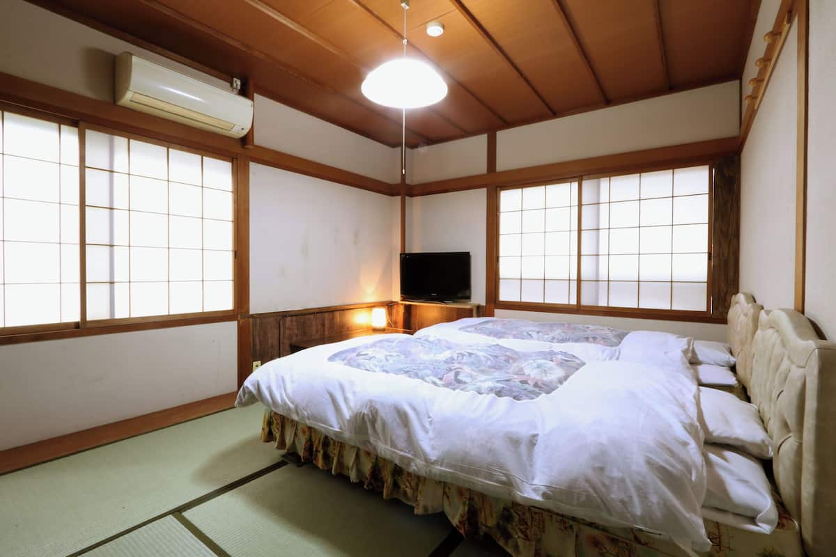 Un dormitorio japonés tradicional con suelo de tatami, una cama grande con colcha estampada con flores, un televisor sobre un soporte de madera y puertas correderas shoji.