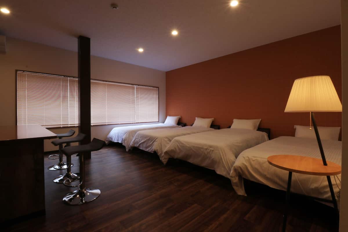 Una habitación de hotel con dos camas, una mesita, un taburete y una ventana con persianas.
