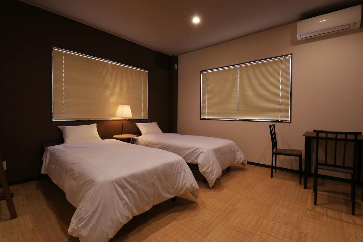 Habitación de hotel con dos camas, un escritorio, una silla y ventanas grandes con persianas.