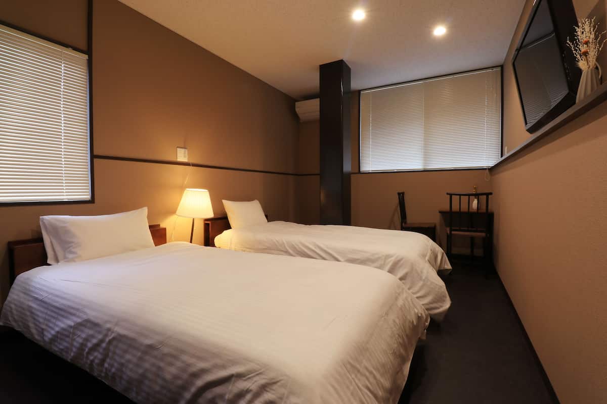 Habitación de hotel con dos camas, una mesita de noche, una lámpara, una silla y un televisor montado en la pared.