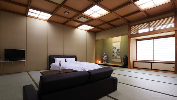 Ein traditioneller japanischer Raum mit Tatami-Boden, einem Bett mit weißer Bettwäsche, einem Fernseher auf einem Regal und einem großen Fenster mit Schiebetüren.