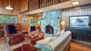 A cozy, rustic interior with a stone fireplace, wooden furniture, and a balcony with a railing. - Cabin With Hot Tub And Private Dock in Bottle Bay (Sagle)