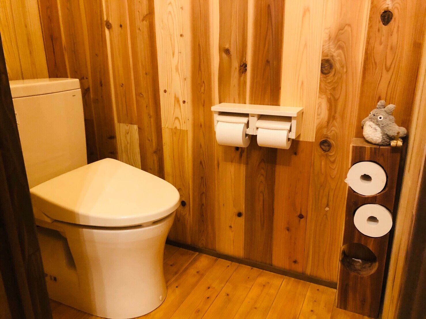 Bagno con rivestimenti in legno, un water, due rotoli di carta igienica e un piccolo ripiano con un peluche.