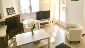Un salon avec une cheminée, un téléviseur, deux fauteuils blancs et une table basse.