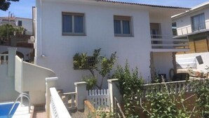 Un immeuble de deux étages, doté d’une façade blanche et d’un toit recouvert de tuiles rouges. On y trouve un petit balcon avec une balustrade en métal et un pot de fleurs.