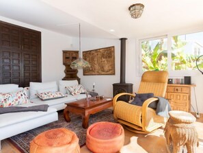 Una sala de estar acogedora con un sofá blanco, un sillón de mimbre y una mesa de centro de madera. Se ve una ventana grande con vistas a zonas verdes.
