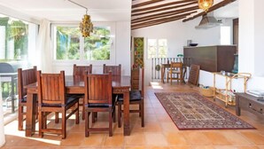 A dining area with wooden chairs and a table, a kitchen with a stove, and a view of greenery through large windows.