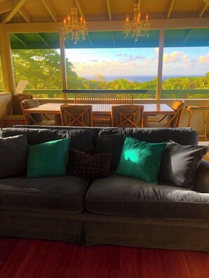 Relax in this serene space with a large sofa, wooden dining table, and a chandelier, offering a view of lush greenery.