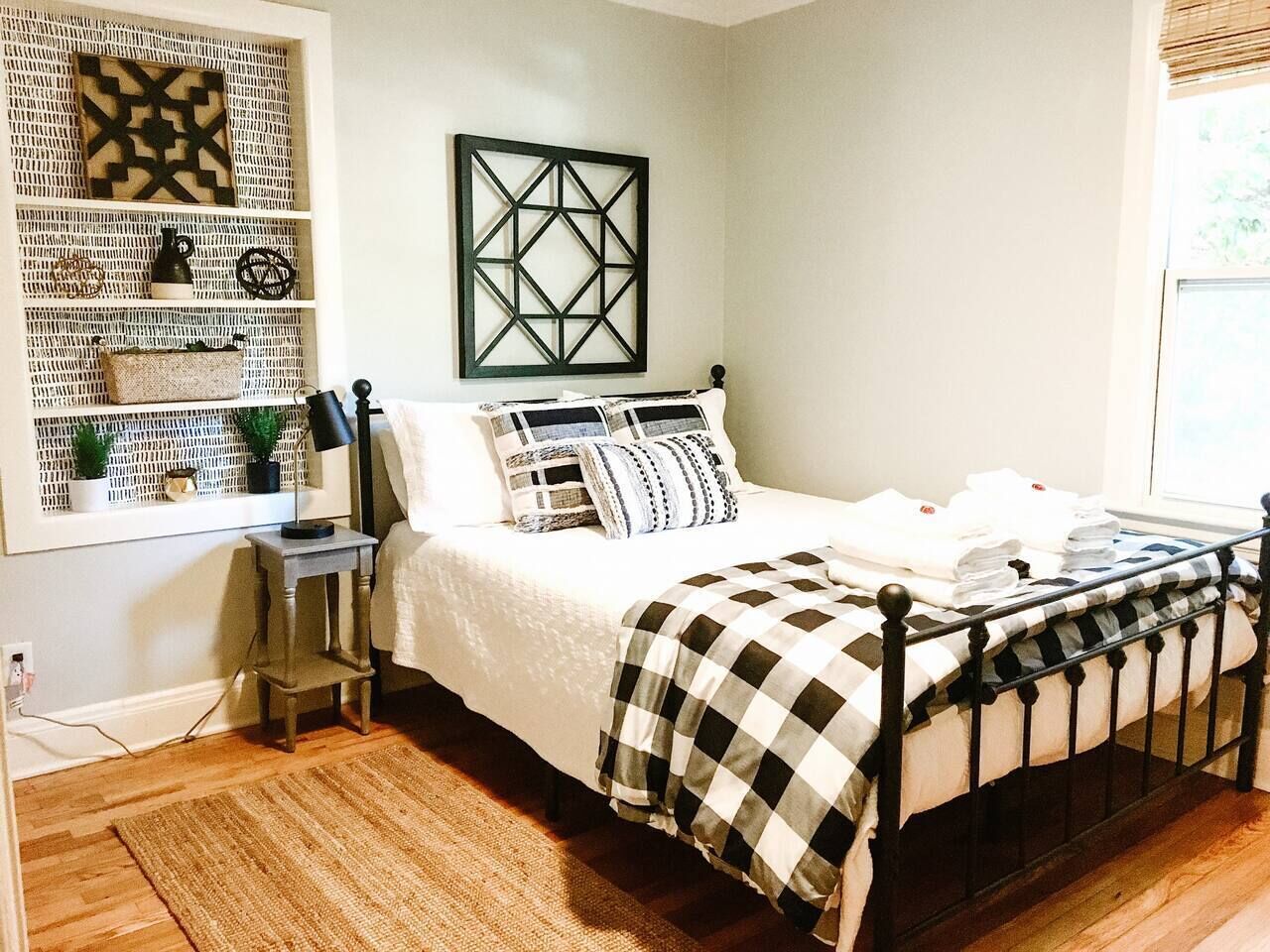 Un dormitorio ordenado con cama, una mesita de noche, un estante en la pared con objetos decorativos y una ventana con persianas.
