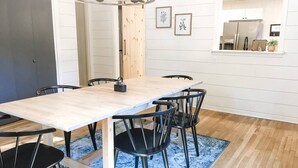 Un coin repas avec une table en bois et des chaises noires, un tapis bleu et une cuisine visible à travers une porte ouverte.