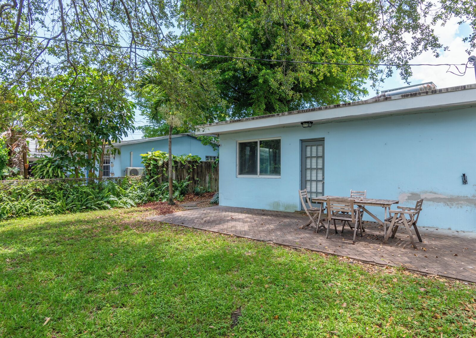 A single-story building with a blue exterior, a patio with a table and chairs, and a lush garden area.