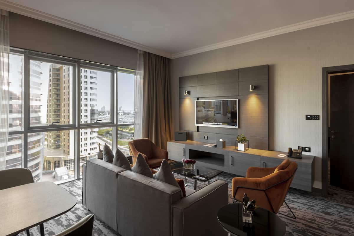 A modern hotel room with a large window, a sofa, a coffee table, and a TV mounted on the wall.