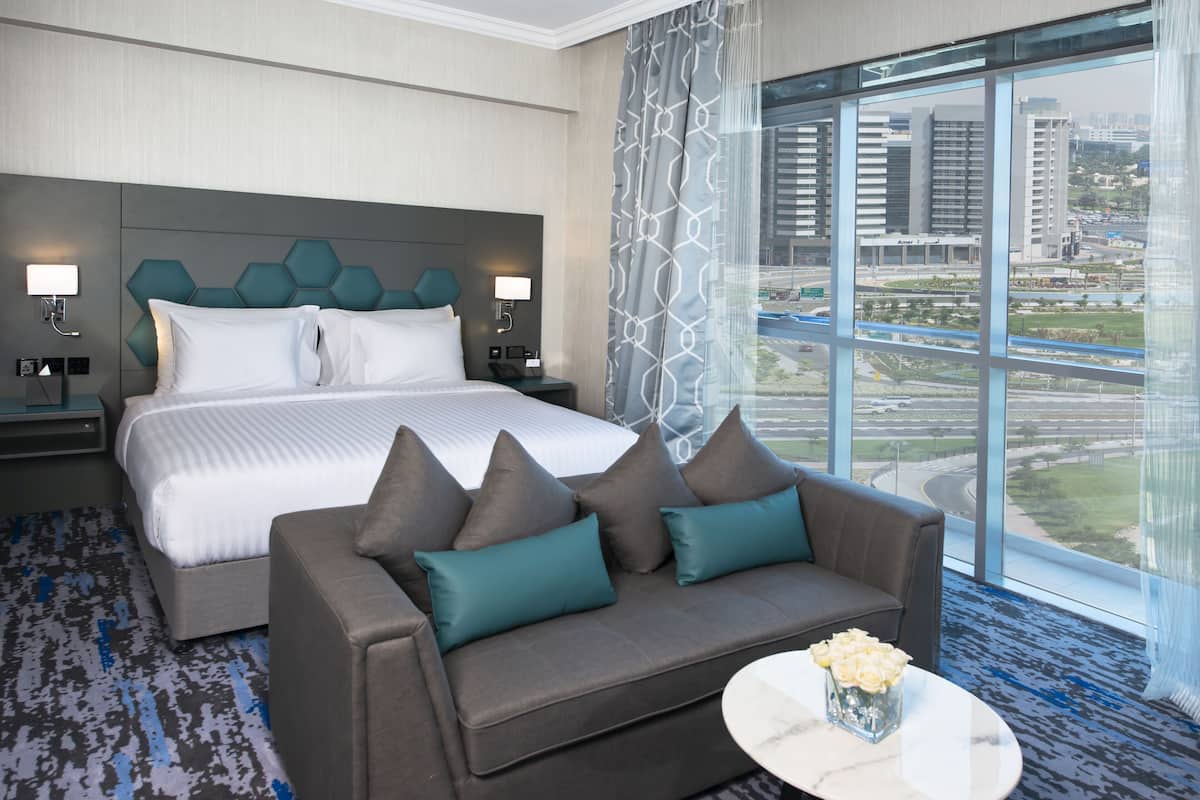 A modern hotel room with a large bed, a grey sofa with teal cushions, a small round table with a vase of flowers, and a city view through large windows.