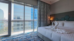 Ein Hotelzimmer mit großem Fenster, einem Bett mit weißer Bettwäsche, einem Kopfteil mit geometrischem Muster und einem Nachttisch mit Lampe.