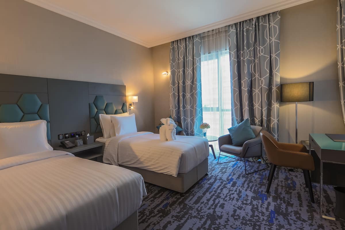 A hotel room with two beds, a desk, a chair, and a large window with patterned curtains.