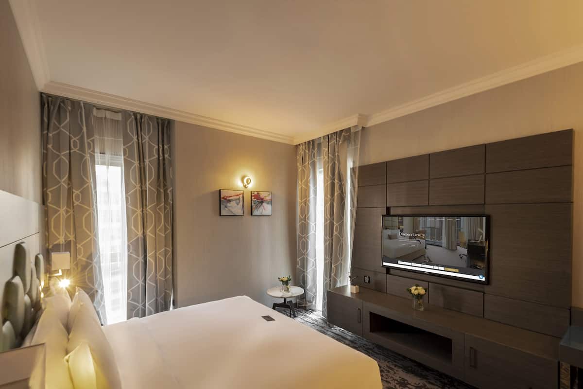 A modern hotel room with a large bed, a flat-screen TV mounted on the wall, a small round table with a vase, and a nightstand with a lamp.
