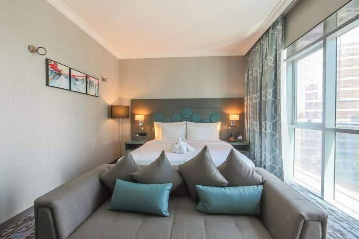 A hotel room with a bed, a sofa with pillows, a nightstand with lamps, and a window with curtains.