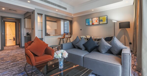 A modern living room with a sofa, armchair, and coffee table, a dining area with chairs, and a kitchen visible through an open door.
