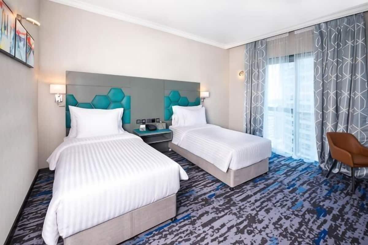 A hotel room with two beds, a nightstand, a chair, and a large window with curtains.