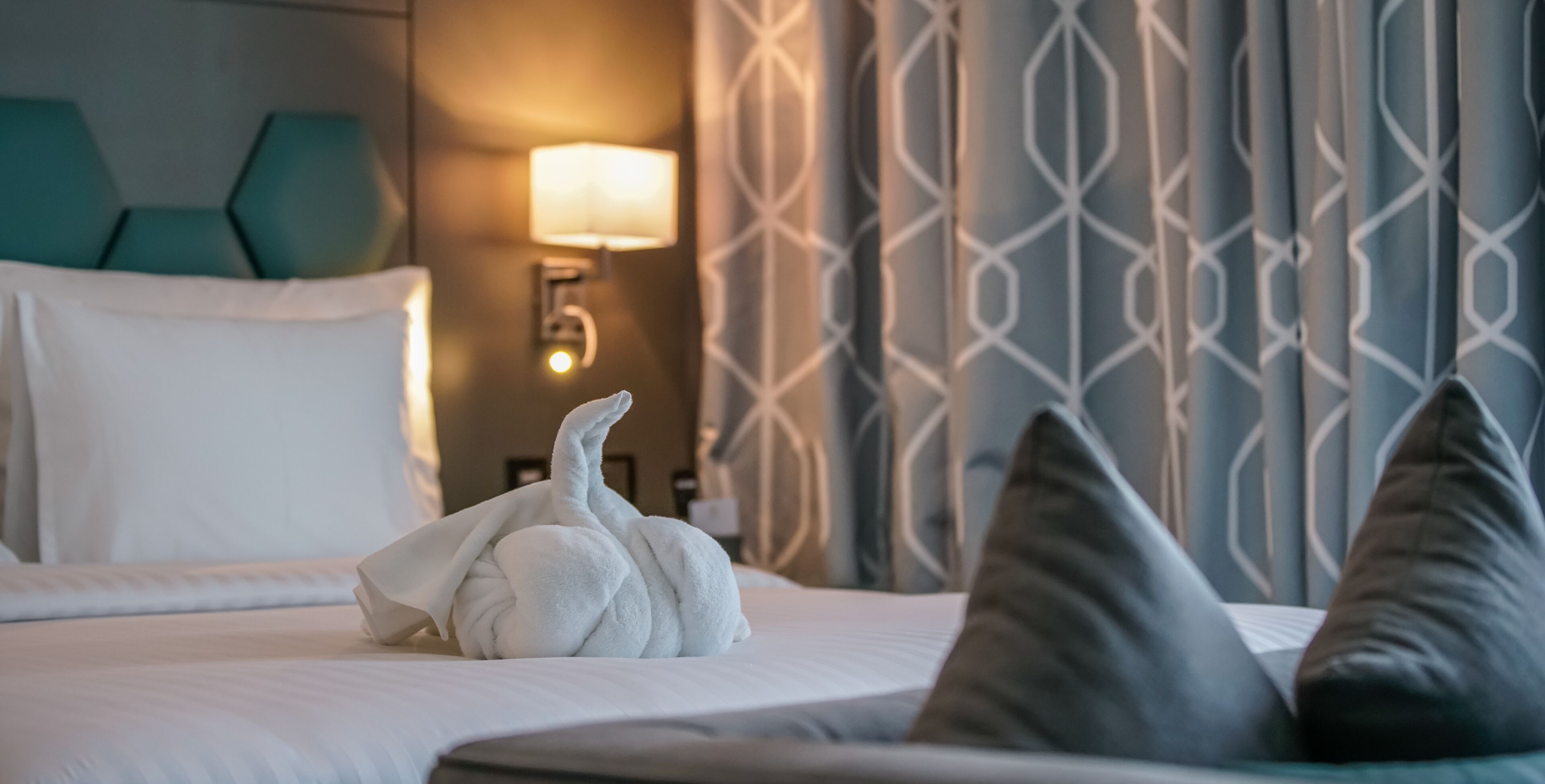 Een net opgemaakt bed met een wit handdoek en kussens, een wandlamp en een gordijn met een patroon.
