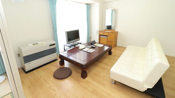 Ein Hotelzimmer mit Holzboden, einem weißen Sofa, einem Holztisch mit Flachbildfernseher, einem Holzkasten und einem Fenster mit Vorhängen.