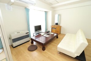 Une chambre d’hôtel avec un plancher en bois, un canapé blanc, une table en bois surmontée d’un téléviseur à écran plat, une armoire en bois et une fenêtre avec des rideaux.