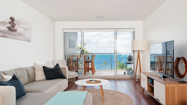 Sala de estar moderna com sofá, mesa de centro e rack para TV. Varanda com vista para o mar visível através de grandes portas de vidro.