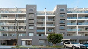 Um edifício residencial com vários andares, varandas, área de estacionamento e uma árvore na frente.