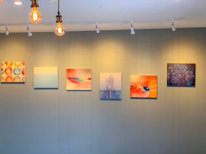 A gallery with six artworks on a wall, illuminated by ceiling lights.