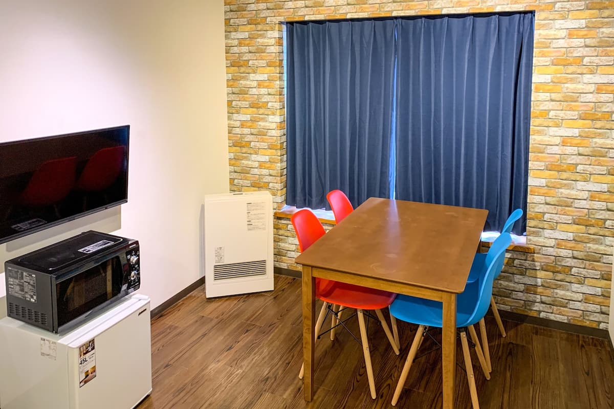 Una habitación con pared de ladrillo, un ventanal con cortinas oscuras, televisor de pantalla plana, microondas, refrigerador pequeño y una mesa de madera con dos sillas.