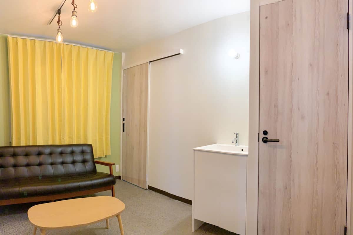 Una habitación con sofá, una mesita, un armario blanco con lavabo y una puerta de madera.