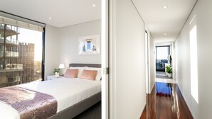 Una camera d'albergo moderna con un letto grande, un comodino con una lampada, una finestra vista sulla città e un corridoio che conduce a un'altra stanza.