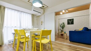 A modern living room with a dining area. It includes a table with yellow chairs, a blue sofa, and a potted plant. - Heights Showa 207 (Sapporo)