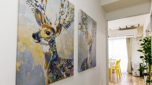 A hallway with two abstract paintings of deer on the wall, a dining area with yellow chairs, and a potted plant. - Heights Showa 207 (Sapporo)