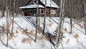 Una cabaña con techo inclinado, rodeada de suelo cubierto de nieve y árboles sin hojas.
