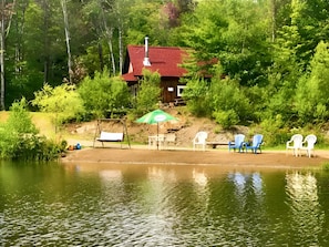 Una cabaña con techo rojo, rodeada de árboles verdes frondosos, cerca de un lago tranquilo con una hamaca y varias sillas Adirondack.
