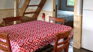 Une table de salle à manger en bois, recouverte d’une nappe à motifs, des chaises en bois, un escalier en bois, un lit et un réfrigérateur blanc.