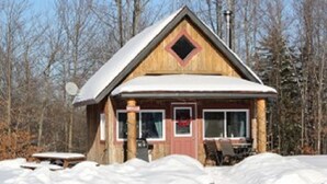 Une cabane en bois avec un toit à deux versants, une porte d’entrée et une petite véranda équipée d’une table et de chaises.