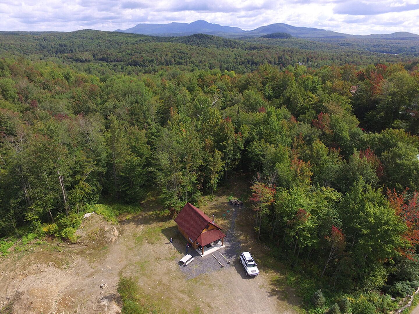 Une vue aérienne d’une cabane entourée d’une forêt dense, avec une chaîne de montagnes en arrière-plan.