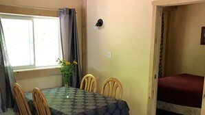 Uma sala de jantar com uma mesa posta para quatro pessoas, cadeiras de madeira, um vaso com flores e uma janela com cortinas.