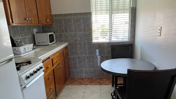 A compact kitchen with a stove, microwave, and cabinets. There's a dining table with chairs and a window with blinds. - Huntington Suites (Huntington Beach)