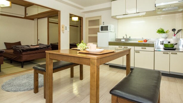A modern kitchen with white cabinets, a microwave, and a sink. There's a dining table with chairs and a bench. - Heights Showa 503 (Sapporo)