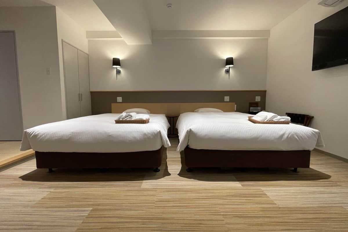 A hotel room with two beds, a wooden floor, a headboard, wall-mounted lights, and a flat-screen TV.