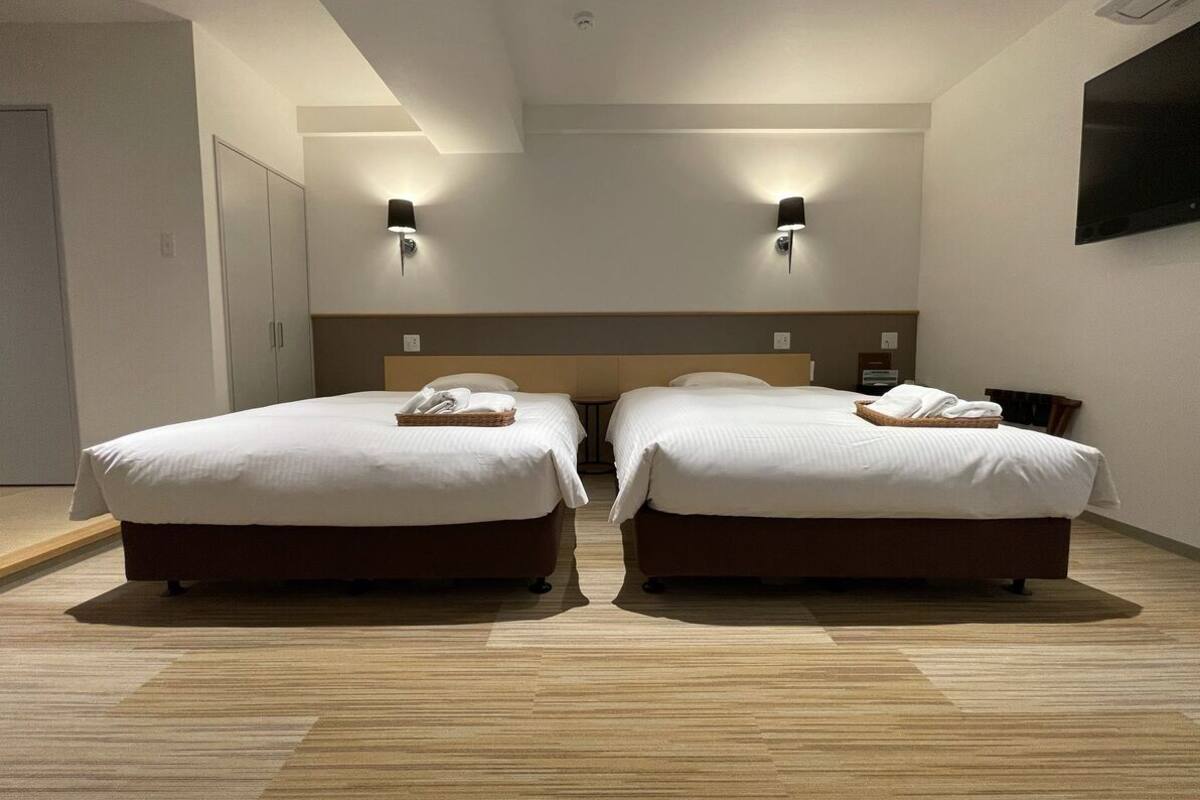 A hotel room with two beds, a wooden floor, a headboard, wall-mounted lights, and a flat-screen TV.