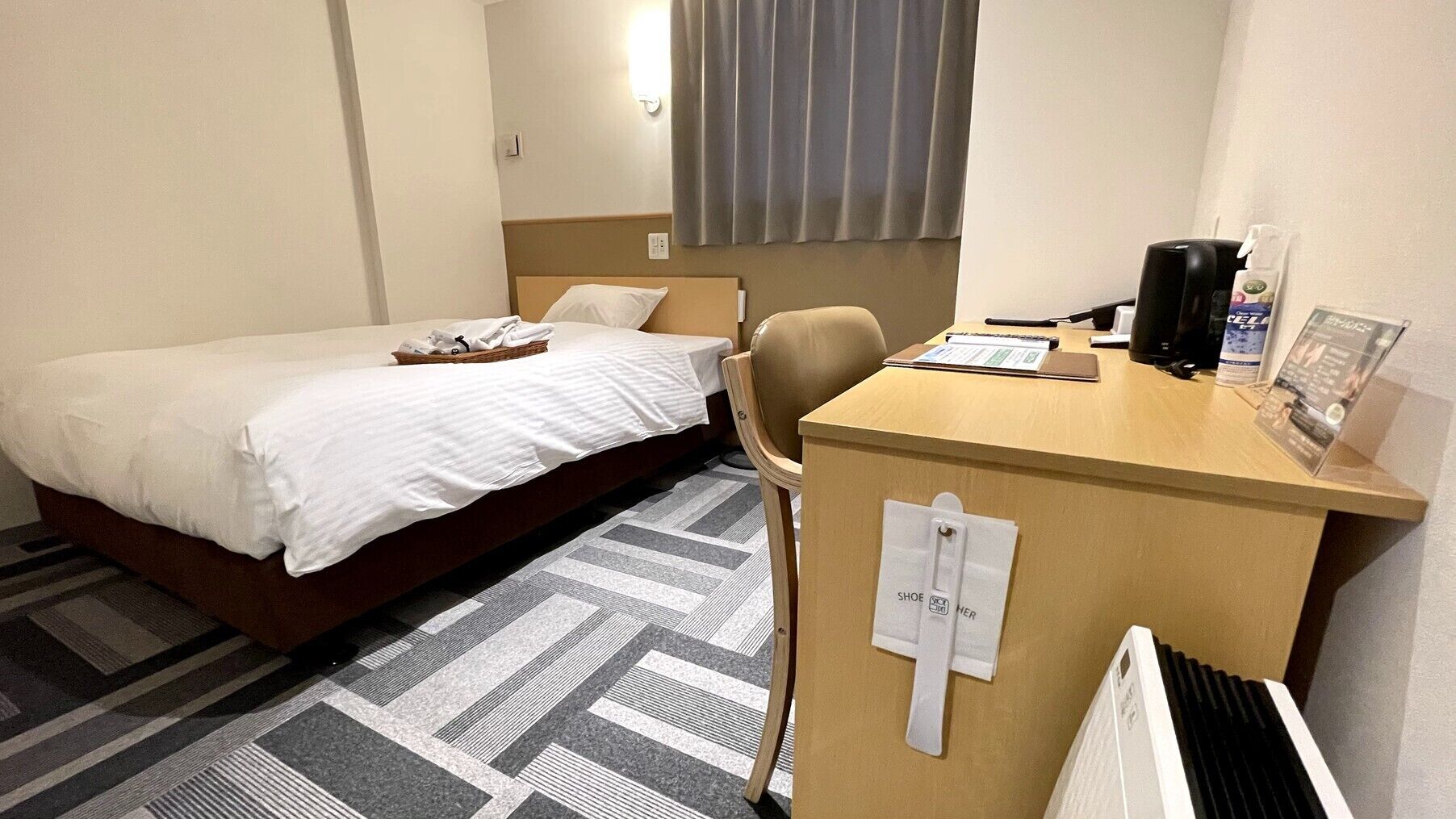 Quarto de hotel com uma cama, uma escrivaninha com cadeira, um radiador e uma janela com cortinas.