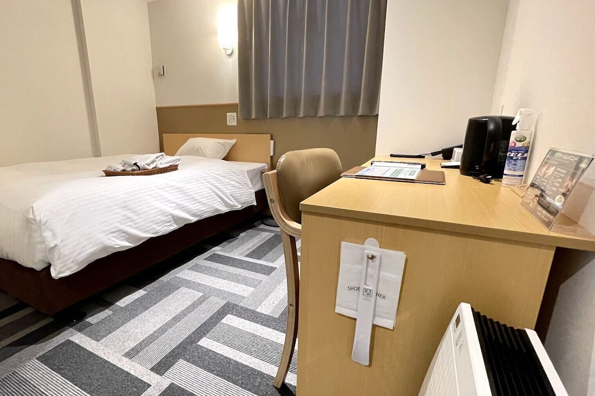 A hotel room with a bed, a desk with a chair, a radiator, and a window with curtains.