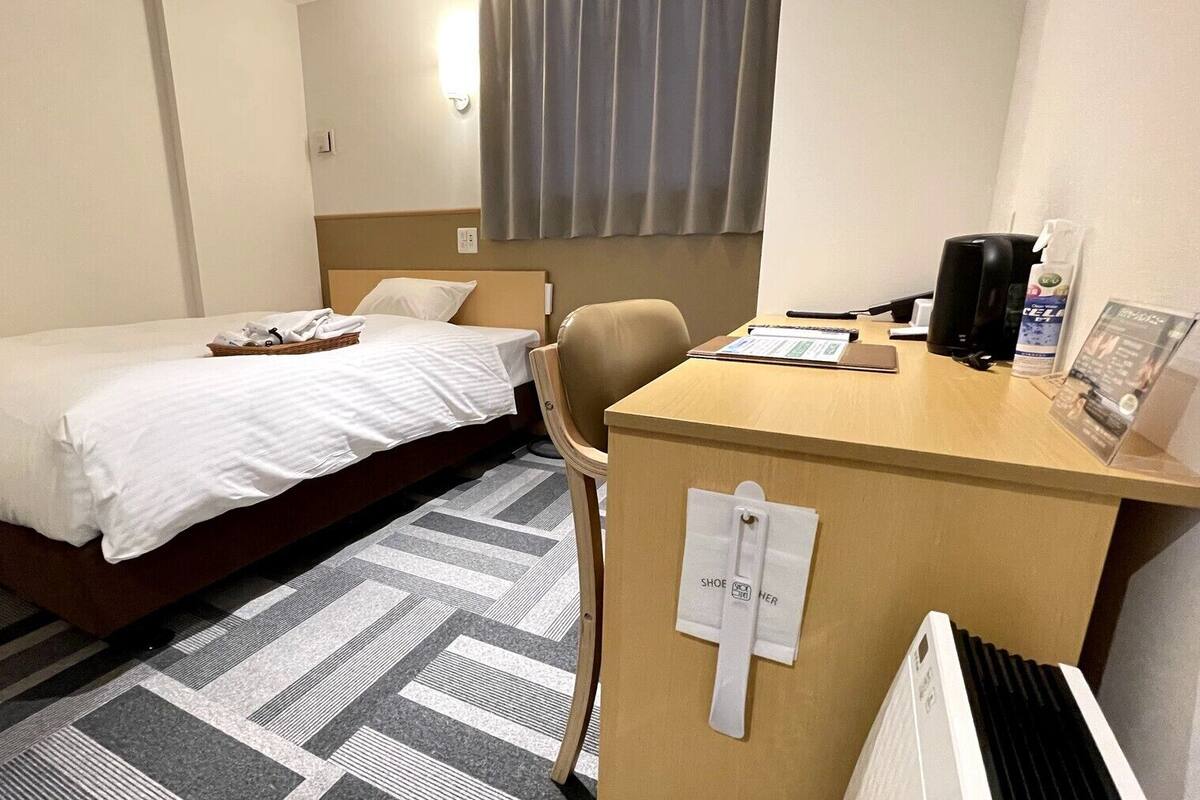 A hotel room with a bed, a desk with a chair, a radiator, and a window with curtains.